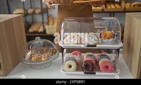 Giovane in una panetteria che tiene in mano una tazza di caffè mentre espone vari dolci e ciambelle sotto contenitori di vetro all'interno Foto Stock