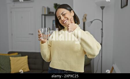 Una giovane donna ispanica sorridente che tiene un bicchiere d'acqua regala un pollice in un accogliente salotto Foto Stock