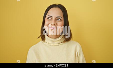 Una giovane donna ispanica sorridente in dolcevita si posa elegantemente su un vibrante sfondo giallo. Foto Stock