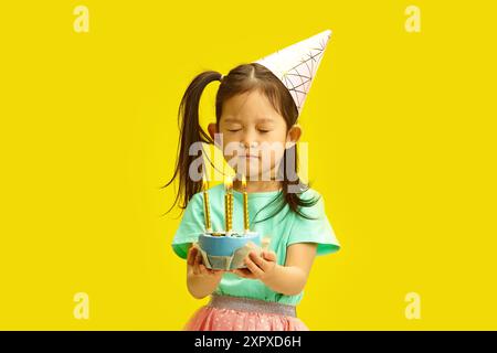 Five Year Old Little Girl with Eyes CLOSED, Making Withday, Hold Cake with lit Candles Standing on Yellow background. Foto Stock