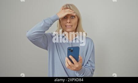 Una donna bionda sorpreso regge uno smartphone su uno sfondo bianco, esprimendo shock o confusione. Foto Stock