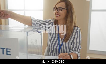 Donna che vota in una stanza elettorale del college, indossa occhiali e camicia a righe, sorride e posiziona un voto in una scatola trasparente con un cordone blu Foto Stock