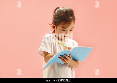 Bambino giapponese che si concentra sulle scritture su copybook, penna in mano, su sfondo rosa. Foto Stock