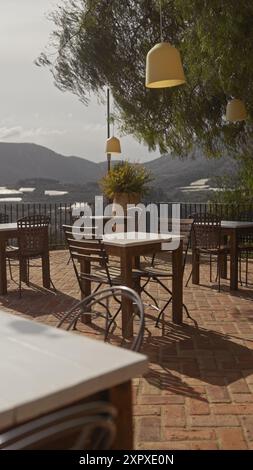 Terrazza all'aperto con sedie e tavoli vuoti che si affacciano sul paesaggio panoramico delle montagne sotto un albero con lampade appese. Foto Stock