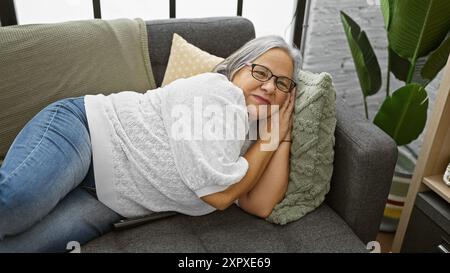 Una donna matura soddisfatta che indossa gli occhiali si appoggia su un divano con un cuscino e una pianta in un accogliente soggiorno. Foto Stock
