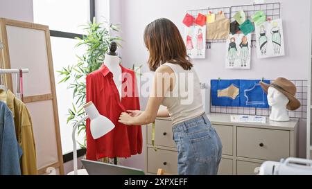 Una giovane stilista lavora su una blusa rossa in un luminoso atelier, circondato da schizzi e campioni di tessuto. Foto Stock