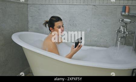 Una giovane donna si rilassa in una vasca da bagno a zampa d'artiglio, usando lo smartphone in mezzo a bolle all'interno di un bagno piastrellato. Foto Stock