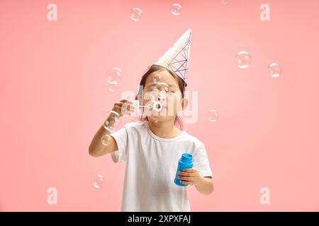 Allegra ragazza di circa 5-6 anni con bolle di sapone Happy Blowing indossate con cappello da festa in carta e maglietta bianca in piedi su sfondo rosa isolato Foto Stock