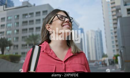 Giovane donna ispanica sorridente mentre cammina su una moderna strada di dubai con grattacieli sullo sfondo. Foto Stock