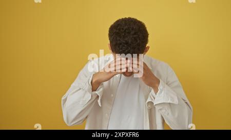 Un giovane stressato con una camicia bianca copre il viso su uno sfondo giallo. Foto Stock
