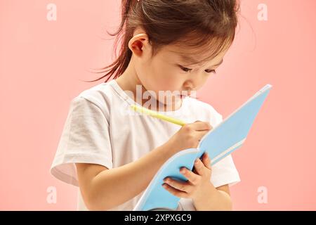 First Grader Girl in abiti casual avvolti scrivendo su notebook in piedi in rosa isolato Foto Stock