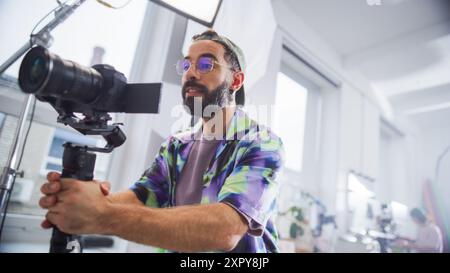 Direttore della fotografia uomo con barba, camicia colorata, lavora con la fotocamera professionale in un ambiente Bright Studio, catturando contenuti coinvolgenti per film o progetti pubblicitari. Foto Stock