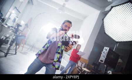 Giovane direttore della fotografia con barba, berretto e camicia a quadri, che filma attivamente in Uno studio professionale, circondato da moderne attrezzature e membri della troupe. Foto Stock