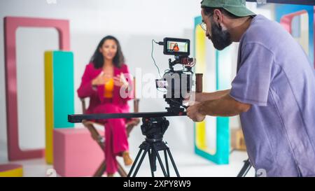 Young Male Cinematographer With Beard, Wearing Cap, Records Vibrant Female Host In Pink Suit During Lively Talk Show Set With Colorful Geometric Shapes, Enhancing Modern Video Production, Storytelling Foto Stock