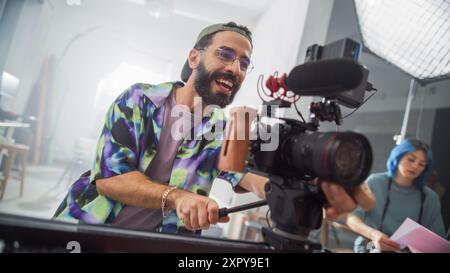 Giovane direttore della fotografia latino maschile con barba, indossa una camicia colorata, lavora con gioia con la fotocamera professionale in ambiente studio, con una collega donna concentrata in background che rivede le note. Foto Stock