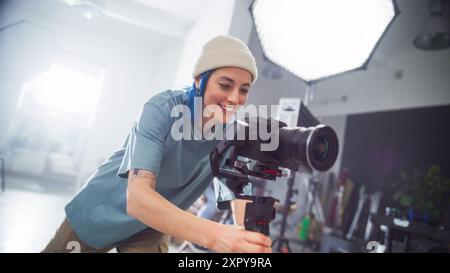 La giovane videografa caucasica adatta la fotocamera professionale in uno studio ben illuminato, catturando l'essenza della moderna produzione cinematografica e la creatività nella produzione pubblicitaria. Foto Stock