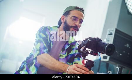 Giovane direttore della fotografia con barba, regolazione delle impostazioni su Una fotocamera moderna durante le riprese di Un film al coperto, indossando Una giacca colorata e occhiali. Creatività e professionalità nel Filmmaking. Foto Stock