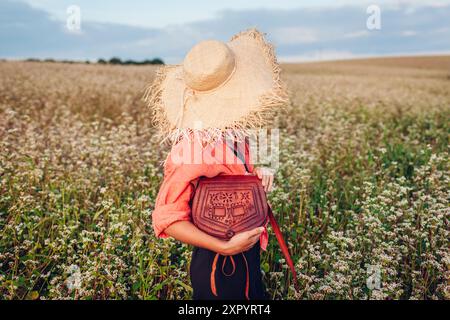 Vista posteriore di una donna elegante che indossa un cappello di paglia nel campo della fibbia in fiore al tramonto tenendo in mano una borsa in pelle. Abbigliamento e accessori alla moda per l'estate Foto Stock