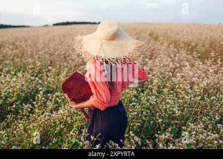 Vista posteriore di una donna elegante che indossa un cappello di paglia nel campo della fibbia in fiore al tramonto tenendo in mano una borsa in pelle. Abbigliamento e accessori alla moda per l'estate Foto Stock