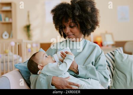 Madre afroamericana che tiene in mano suo figlio e gli dà da mangiare mentre il bambino cerca di tenere il biberon da solo Foto Stock