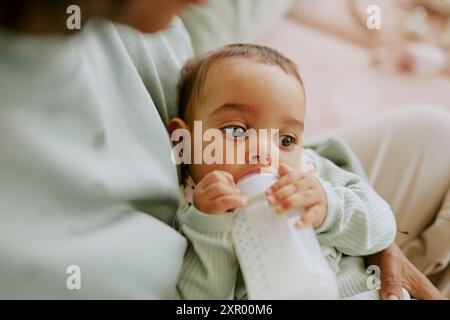 Primo piano medio di un neonato maschio che tiene il biberon con il latte in polvere e che mangia da solo mentre la madre lo tiene in braccio Foto Stock