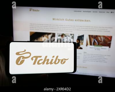 Persona che possiede uno smartphone con il logo della società tedesca di vendita al dettaglio di caffè Tchibo GmbH davanti al sito web. Mettere a fuoco il display del telefono. Foto Stock