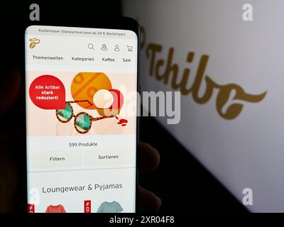 Persona che detiene un cellulare con la pagina web della società tedesca di vendita al dettaglio di caffè Tchibo GmbH davanti al logo aziendale. Messa a fuoco al centro del display del telefono. Foto Stock