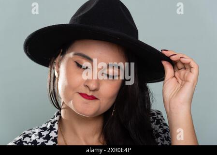 Ritratto ravvicinato di una bella donna sorridente con i capelli neri che indossa un cappello nero in posa per la foto in un abito bianco e nero. Isolato su verde chiaro b Foto Stock