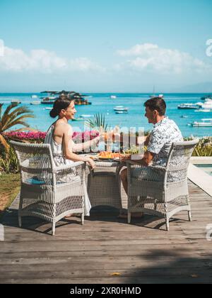Una coppia si diverte con un drink e un pasto in riva al mare con le barche sullo sfondo in una giornata di sole. Foto Stock