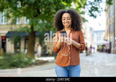 Una donna felice sta usando il suo smartphone mentre cammina lungo una strada urbana. Indossa le cuffie e sembra che stia godendo il suo tempo all'aperto. La scena cattura uno stile di vita moderno e rilassato in città. Foto Stock