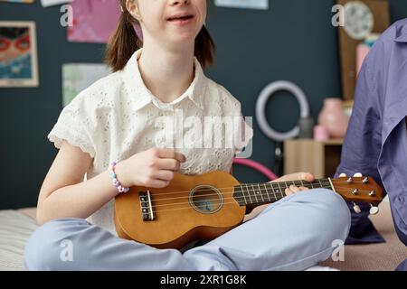 Giovane ragazza con un'espressione gioiosa seduta nell'interno di casa, tenendo in mano un ukulele mentre è circondata da un arredamento colorato e da morbidi cuscini, mostrando entusiasmo per la musica Foto Stock