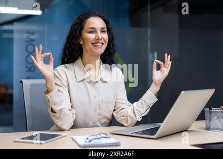 Donna d'affari che pratica meditazione sulla consapevolezza alla scrivania promuovendo la salute mentale sul posto di lavoro. Una donna siede alla scrivania con un computer portatile e un telefono, sorridendo e mostrando relax Foto Stock