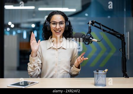 Donna sorridente che indossa le cuffie per registrare podcast con microfono in studio. Persona che gestiva le mani durante la trasmissione. Il telefono tablet sulla scrivania indica l'uso della tecnologia. Foto Stock