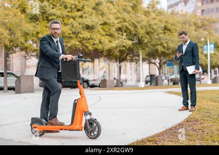 Concetto di trasporto ecologico. Uomo d'affari in tuta a bordo di uno scooter elettrico durante una riunione di lavoro. Uomo d'affari felice con scooter all'aperto Foto Stock