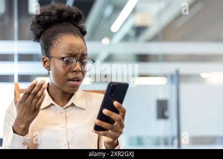 Giovane donna d'affari afroamericana all'interno dell'ufficio che guarda sorpreso al telefono. Indossando occhiali e camicia bianca, sembra confusa o scioccata dalle informazioni che vede sul suo telefono. Foto Stock