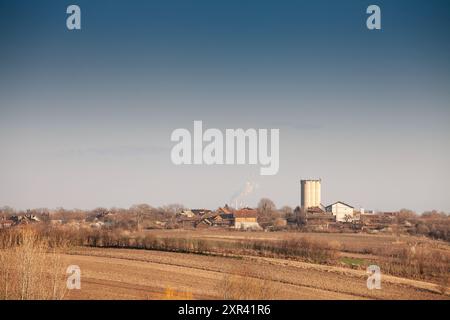 Questa vista panoramica cattura un piccolo villaggio in Vojvodina, Serbia, sullo sfondo di ampi campi agricoli. La scena riflette il rur Foto Stock