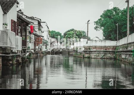 Un canale attraversa un tradizionale villaggio cinese con pareti bianche, un ponte di pietra e lussureggianti alberi verdi. Foto Stock