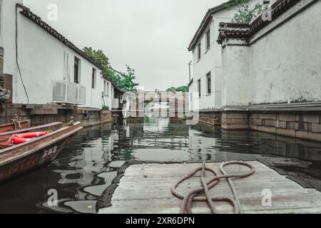 Una barca di legno è legata ad un molo di legno intempestivo in uno stretto canale. Il canale scorre tra due edifici bianchi con tetti neri ornati e una pietra Foto Stock