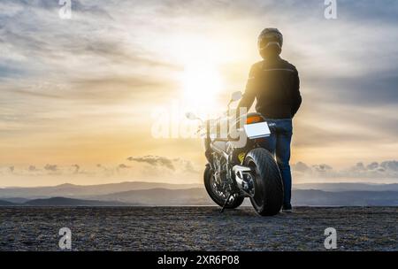 motociclista in piedi accanto alla motocicletta, guardando il tramonto - spazio copia. Foto Stock