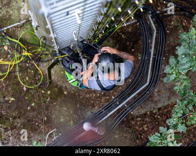 Il tecnico delle telecomunicazioni sta lavorando in quota, indossando un'imbracatura di sicurezza e un casco, installando cavi in fibra ottica su una struttura metallica Foto Stock