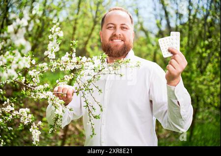Uomo allergico che soffre di allergia stagionale in primavera. Bell'uomo che tiene in mano una confezione di pillole, applica farmaci allergici, posa in giardino fiorito in primavera. Concetto di farmaco antistaminico Foto Stock