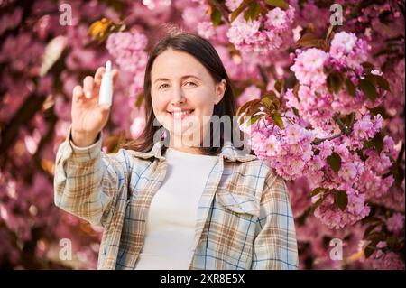 Donna allergica usando gocce nasali mediche, soffre di allergia stagionale nel giardino fiorito. Donna sorridente che tiene uno spray nasale vicino a un albero in fiore. Concetto di allergia primaverile. Foto Stock