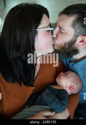 I genitori si baciano mentre tengono in braccio il neonato con un vestito grigio. Foto Stock