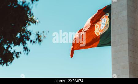 La bandiera portoghese ondeggia orgogliosamente contro un cielo azzurro, parzialmente incorniciato da un ramo d'albero sulla sinistra. I vivaci colori rosso e verde si distinguono, Foto Stock