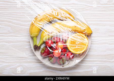 Frutta e frutti di bosco assortiti su piastra bianca avvolta in pellicola trasparente di plastica vista dall'alto su sfondo rustico bianco in legno Foto Stock