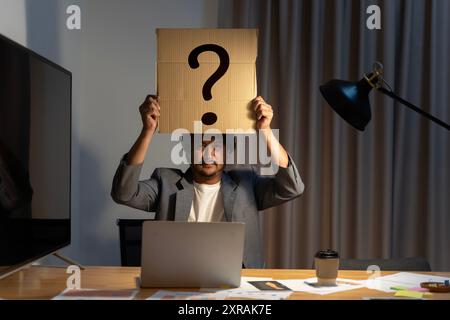 Giovane con un cartello con la domanda su una carta che copre la faccia. Dilemma del dipendente con punto interrogativo su carta bianca. Confuso, pensiero, brainstorming o dec Foto Stock