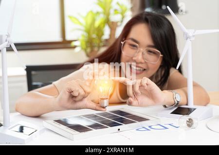 Donna che tiene la lampadina su un tavolo bianco con modello di turbina eolica, cella solare e casa a blocchi sul tavolo da lavoro a casa che pianifica di risparmiare energia. Verde en Foto Stock