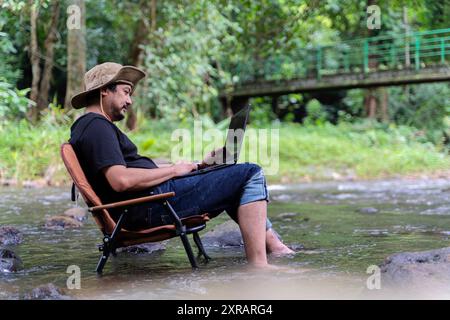 Bell'uomo asiatico barbuto in età lavorativa seduto su una sedia da campeggio e che lavora su un computer portatile vicino al ruscello in mezzo al parco nella foresta all'aperto da solo felicemente Foto Stock