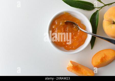 Dettaglio di un piccolo piatto con marmellata di pesca su un banco bianco con frutta e foglie intorno ad esso. Vista dall'alto. Foto Stock