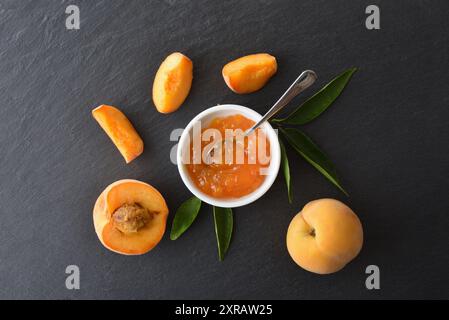 Dettaglio di un piccolo piatto con marmellata di pesca su un banco bianco con frutta e foglie intorno ad esso. Vista dall'alto. Foto Stock